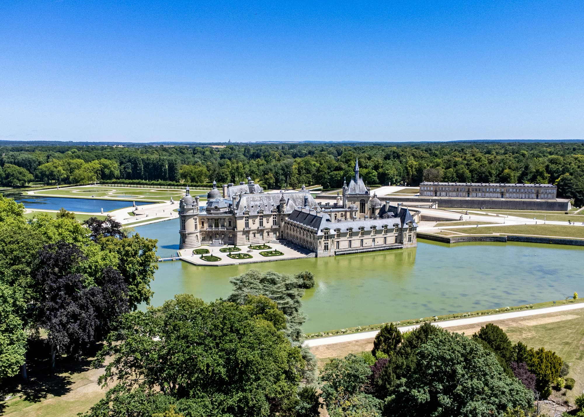 The Château de Chantilly from the air, ringed by green water with the Le Nôtre park and forest behind it