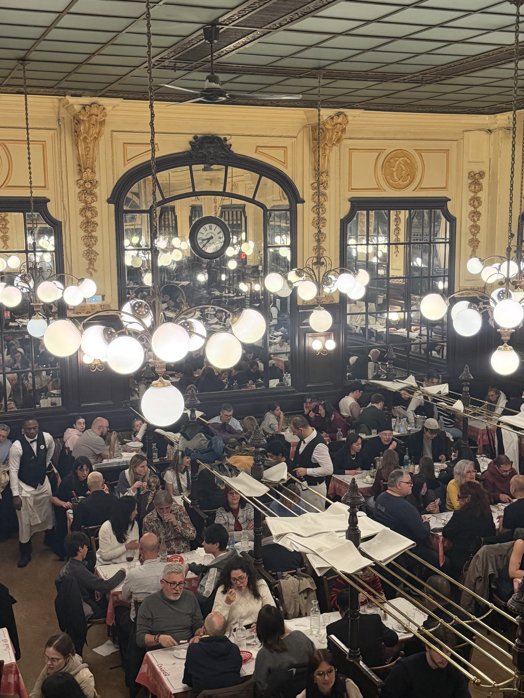 The dining room at Bouillon Chartier from the balcony, globe lamps and gilded mirrors above rows of full tables, waiters in waistcoats between them